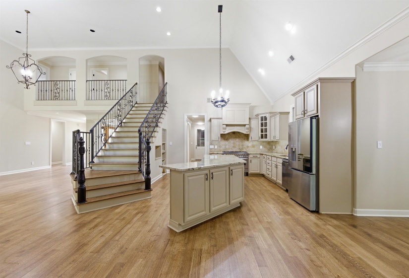 Open-concept living area featuring wood-finish flooring, a grand staircase with wrought iron balusters, a kitchen island with stone countertop, stainless steel appliances, and a vaulted ceiling