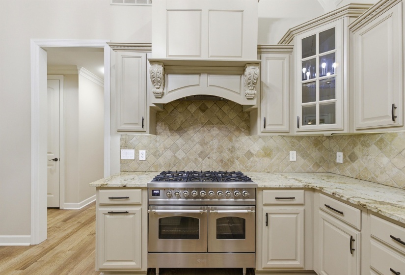 Gourmet kitchen featuring a professional-grade stainless steel range with double ovens, a decorative range hood, extensive light-toned cabinetry with glass-front accents, granite countertops, and a diamond-patterned tile backsplash