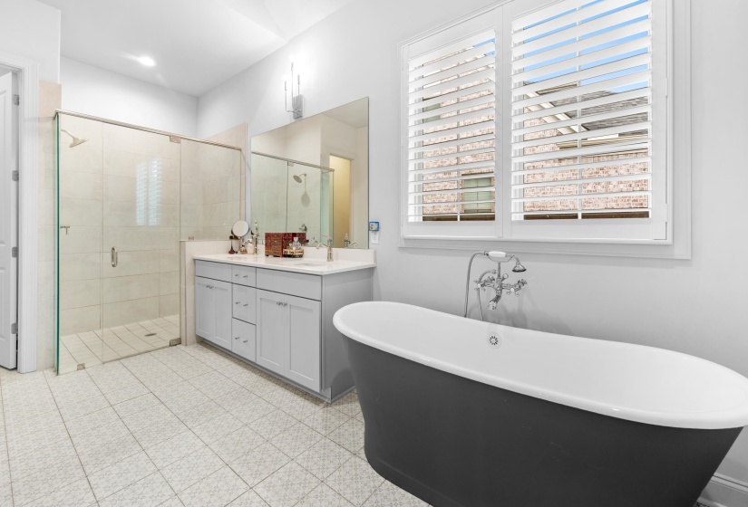 Spacious bathroom featuring a freestanding soaking tub, a dual vanity with a light gray cabinet, a glass-enclosed shower, white tile flooring with a subtle pattern, and window shutters
