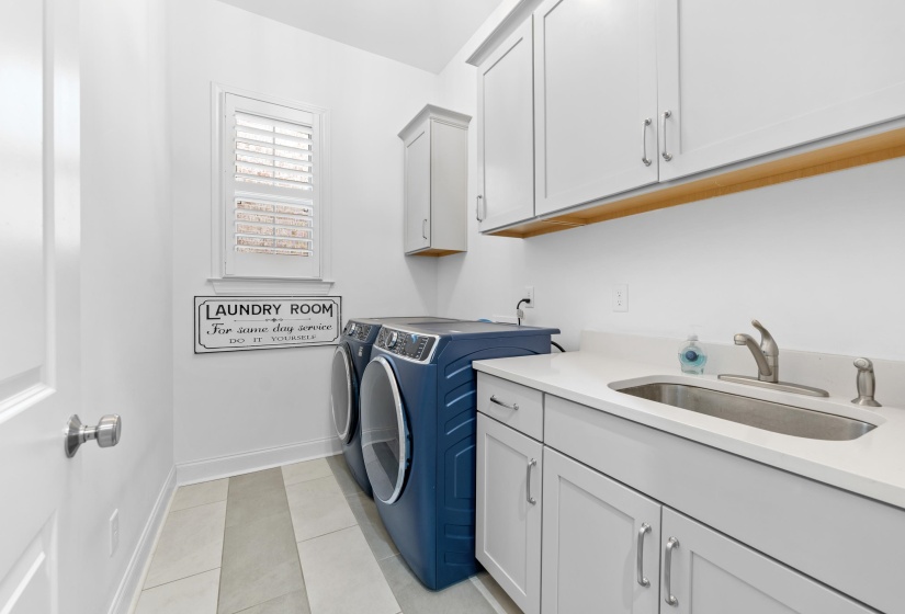 Laundry room featuring a single window with plantation shutters, white shaker-style cabinetry, a built-in sink with a countertop, and a wood-finish shelf
