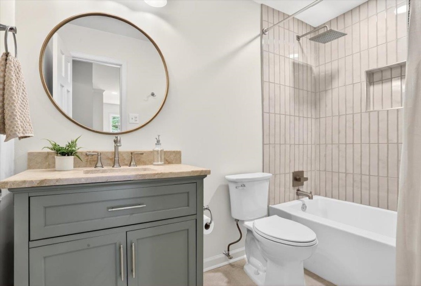 Bathroom featuring a gray vanity with a stone countertop, a circular framed mirror, a white toilet, and a tub with a tiled shower surround and a rainfall showerhead