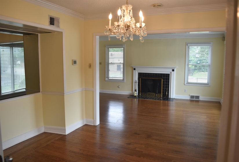 Hardwood flooring extends throughout this open living area featuring a decorative chandelier, a fireplace with a black tile surround and white mantel, and multiple windows with blinds