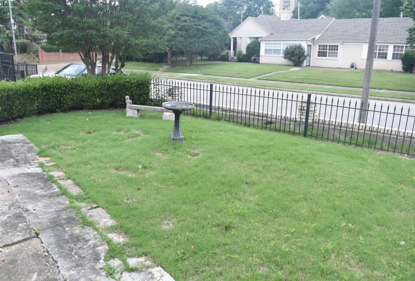Front yard featuring a black wrought iron fence, mature shrubbery, green lawn, and concrete pathway