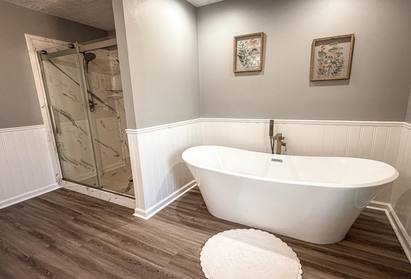 Freestanding soaking tub with a floor-mounted faucet, wood-finish flooring, wainscoting, and a glass-enclosed shower featuring a marble-look surround