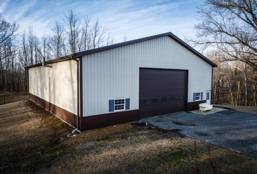 Two-tone metal building featuring a large brown overhead door with windows, two white framed windows with blue shutters, a white entry door, and a brown metal roof
