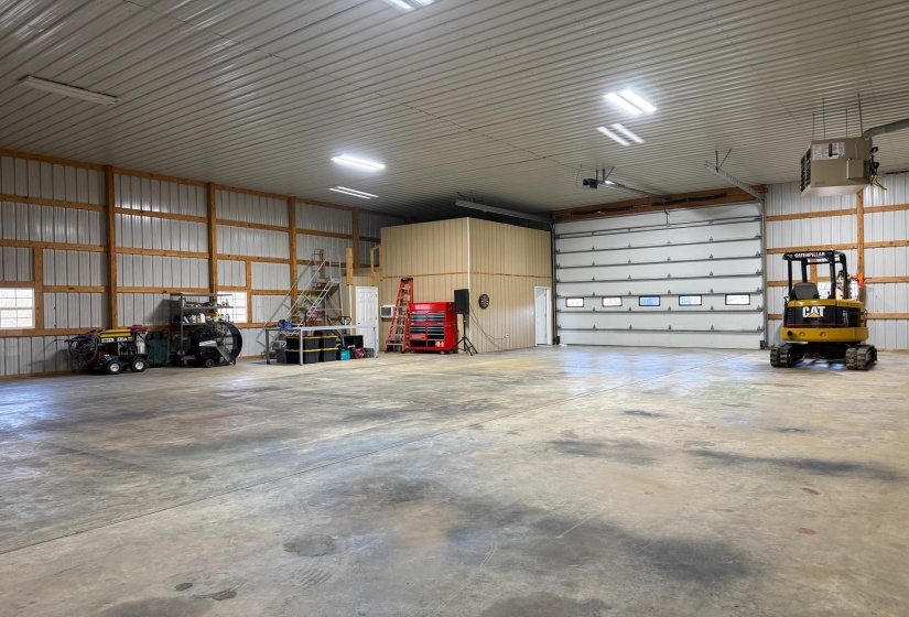 Spacious interior featuring a concrete floor, high ceilings with exposed beams, multiple fluorescent lighting fixtures, and a large overhead door with windows