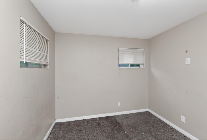 Neutral-toned walls with baseboard trim, featuring two windows with horizontal blinds and a gray carpeted floor