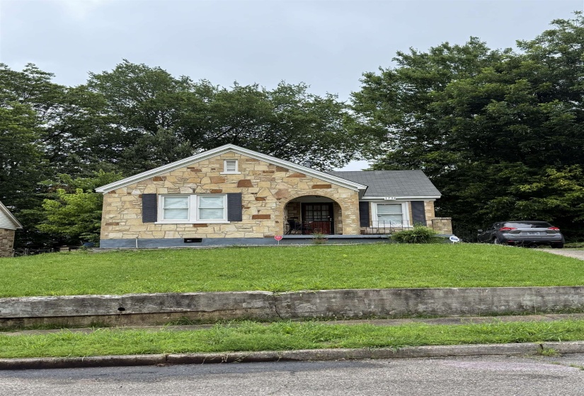 Stone façade residence featuring a prominent arched entry, dark window shutters, a gabled roofline, and a sloped front lawn