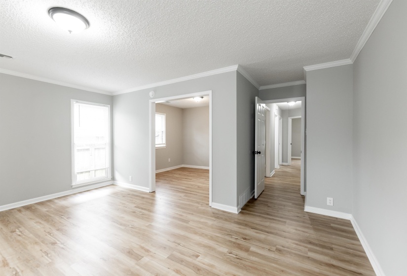 Spacious interior showcasing wood-finish flooring, light gray wall tones, white trim, a flush mount ceiling light, and an open doorway to an additional room