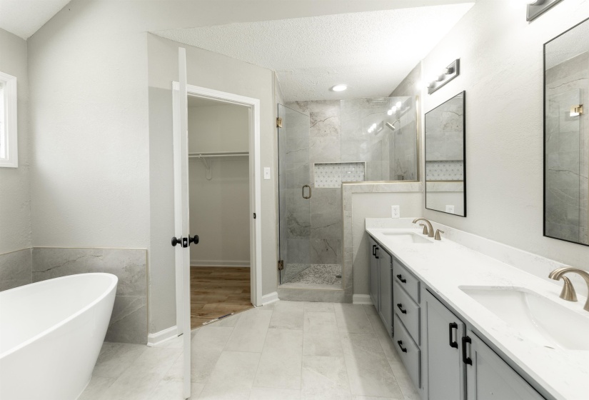 Spacious bathroom featuring a freestanding soaking tub, a double vanity with light gray cabinetry and white countertops, a walk-in shower with glass enclosure and tiled walls, and light tile flooring