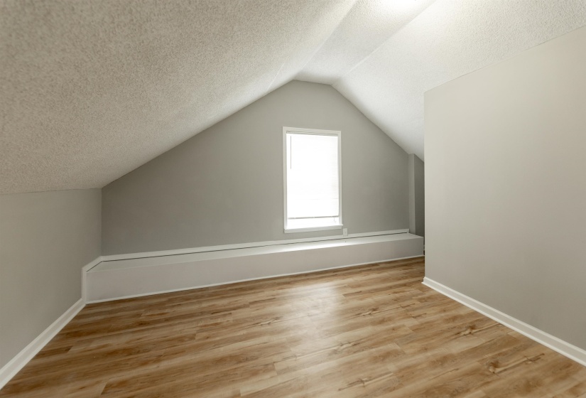 Attic space featuring wood-finish flooring, light grey painted walls, white baseboards, and a window with white trim