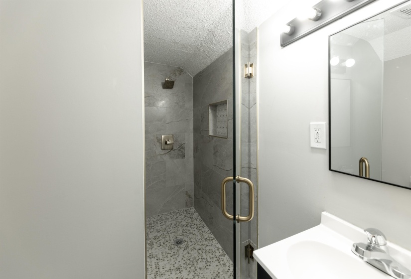 Walk-in shower featuring a glass enclosure with gold-tone hardware, large format grey tile walls, a built-in shower niche, and a contrasting mosaic tile floor