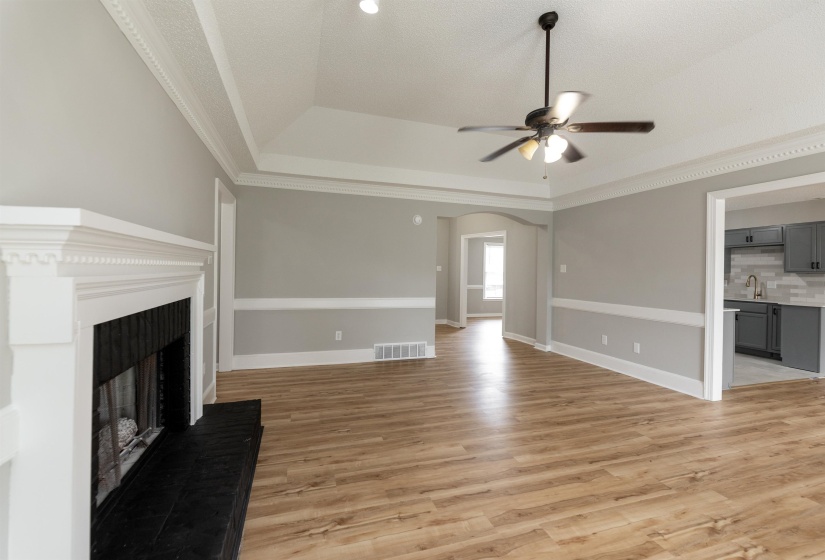 Spacious room featuring wood-finish flooring, a white mantel fireplace with a black hearth, tray ceiling with a ceiling fan, and crown molding