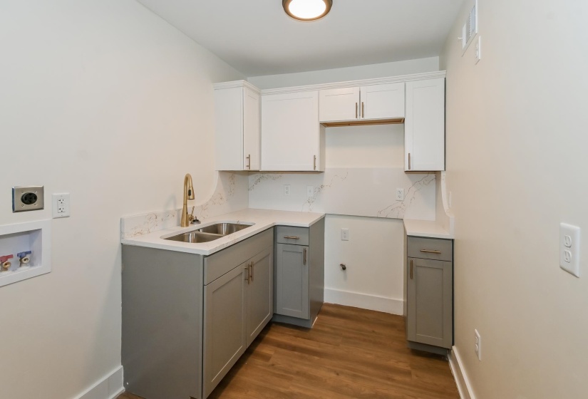 Laundry area featuring wood-finish flooring, two-tone cabinetry with upper white and lower gray cabinets, a double basin sink with a gold-tone faucet, and white countertops with a matching backsplash