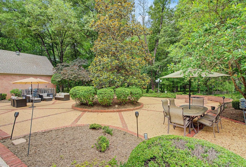 Expansive outdoor living area featuring a patterned paver patio with brick accents, mature trees, landscaped planting beds, and a built-in fire pit