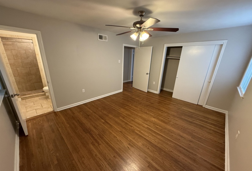 Hardwood flooring in a bright room featuring neutral wall paint, a ceiling fan with integrated lighting, a sliding door closet, and an attached bathroom with tiled flooring and shower surround