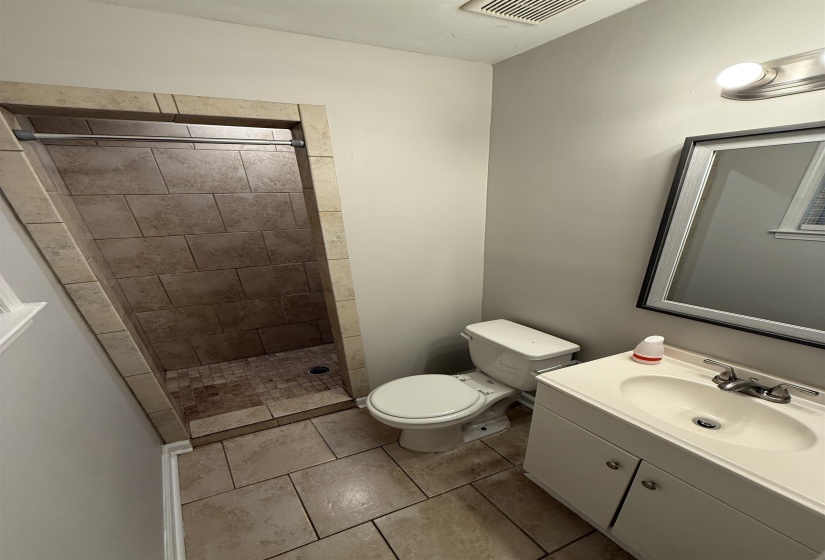 Bathroom featuring a walk-in shower with tile surround, a single vanity with an integrated sink, a toilet, and light-toned tile flooring