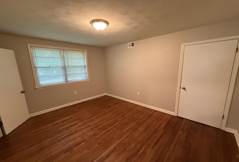 Spacious room featuring rich wood-finish flooring, light tan wall paint, white trim, and a prominent window