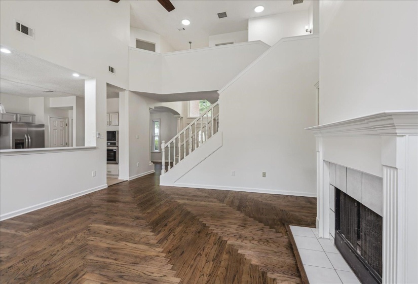 Living area featuring wood-finish flooring in a chevron pattern, a white mantel fireplace with a tile hearth, and a staircase with white balusters