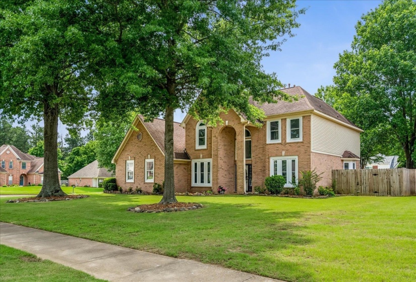 Traditional-style residence featuring a brick facade with light-toned siding accents, a prominent arched entry, multiple white-trimmed windows, a gabled roofline, and mature trees framing the expansive front lawn