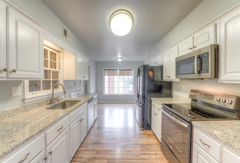 Galley kitchen featuring white cabinetry, light-colored countertops, stainless steel appliances, wood-finish flooring, and a large window providing natural light