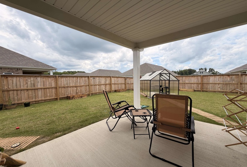 Expansive backyard featuring a covered concrete patio, wood privacy fencing, a glass-paneled greenhouse structure, and a lush green lawn
