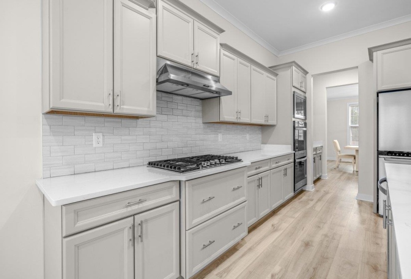 Contemporary kitchen featuring light wood-finish flooring, white shaker-style cabinetry, a tile backsplash, and stainless steel appliances including a gas cooktop and double wall ovens