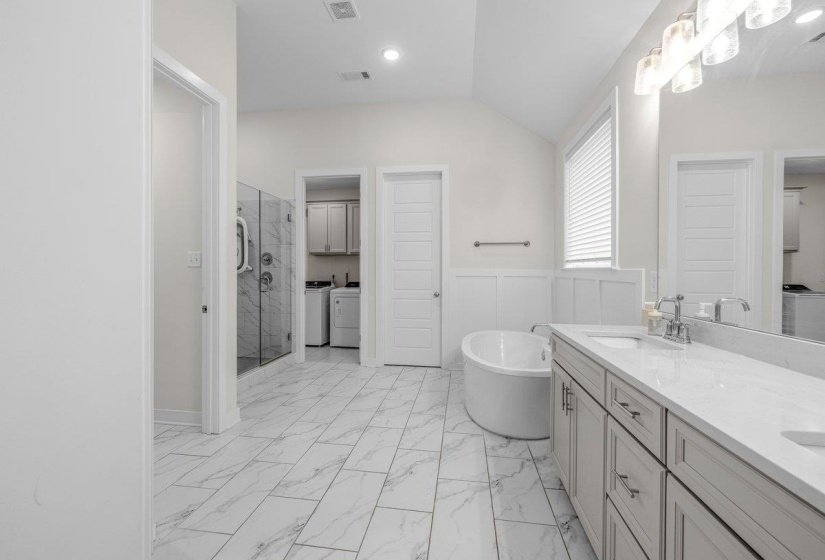 Spacious bathroom featuring white rectangular tile flooring, a dual vanity with light gray cabinetry and a white countertop, a freestanding soaking tub, and a separate glass-enclosed shower