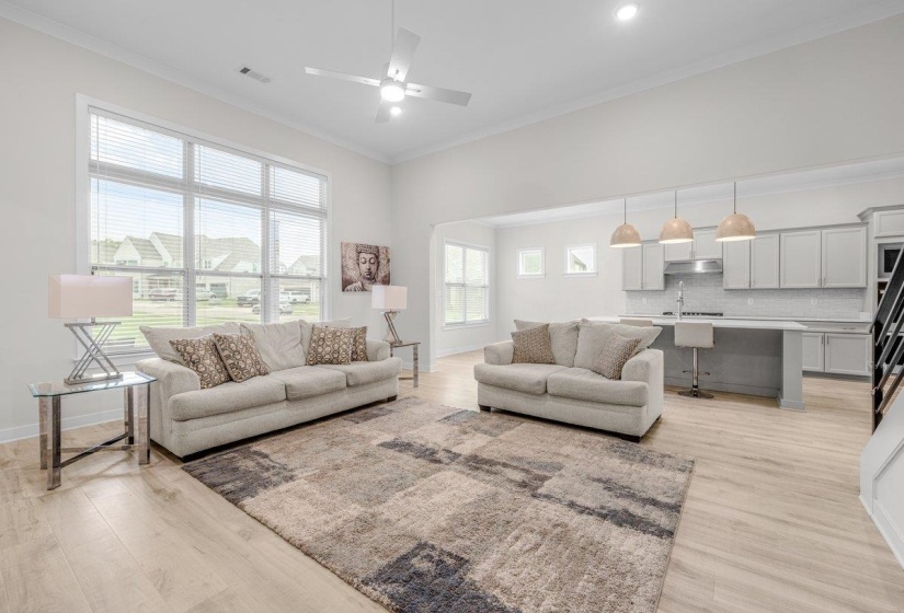 Open-concept living space featuring high ceilings with a ceiling fan, abundant natural light through large windows, light wood-finish flooring, a kitchen island with seating, and light-toned cabinetry