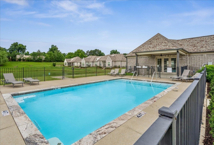 Outdoor swimming pool with integrated steps, surrounded by a concrete deck with stone-finish coping, a black metal fence, and an adjacent brick building with a covered patio
