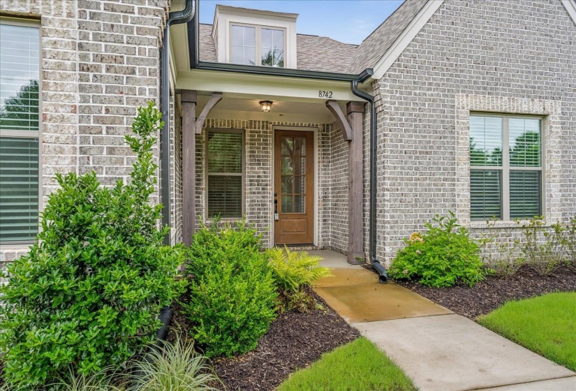 Brick facade entry featuring a covered porch with exposed timber columns, a paneled wood-finish front door with glass inserts, and a concrete walkway
