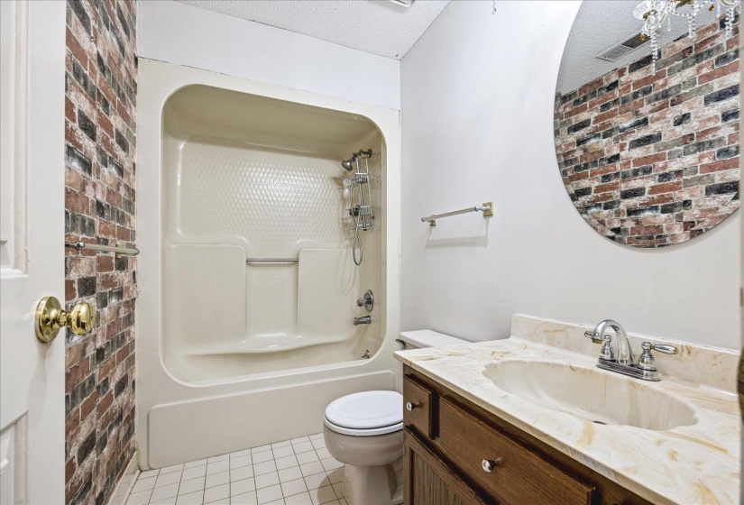 Bathroom featuring a shower/tub combination, brick-patterned accent wall, wood-finish vanity with a single basin, light-toned countertop, and a round wall mirror