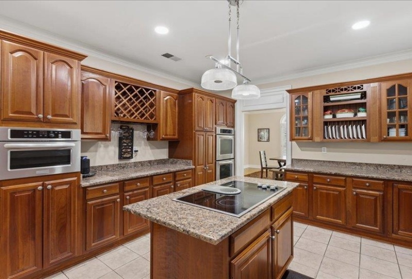 Kitchen featuring a central island with a built-in cooktop, extensive wood cabinetry, granite-finish countertops, stainless steel wall oven and microwave, and ceramic tile flooring