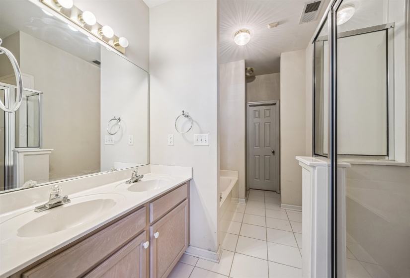 Bathroom featuring a dual-sink vanity with light wood-finish cabinetry, a large wall-mounted mirror, a separate glass-enclosed shower, a built-in bathtub, and ceramic tile flooring