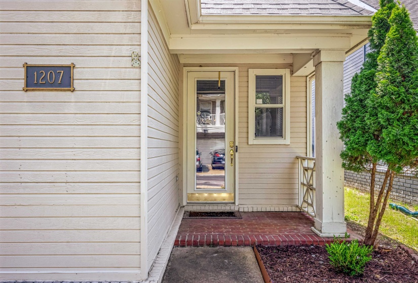 Welcoming front entry featuring light-colored siding, a covered porch with brick flooring, a full-lite entry door, and a decorative house number plaque