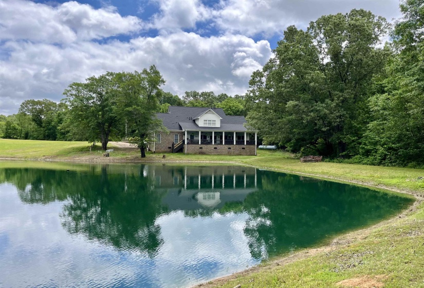 Expansive pond with a lush green perimeter, complemented by a home featuring a covered porch, brick foundation, and a dark shingle roof, all surrounded by mature trees and natural landscaping