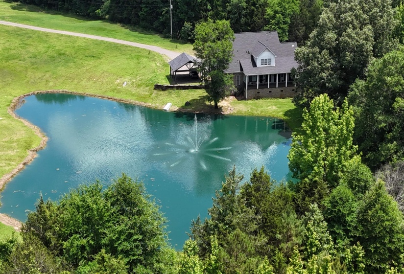 Expansive property featuring a pond with a fountain, a large single-story residence with a covered porch, an additional detached covered structure, mature trees, and a gravel driveway