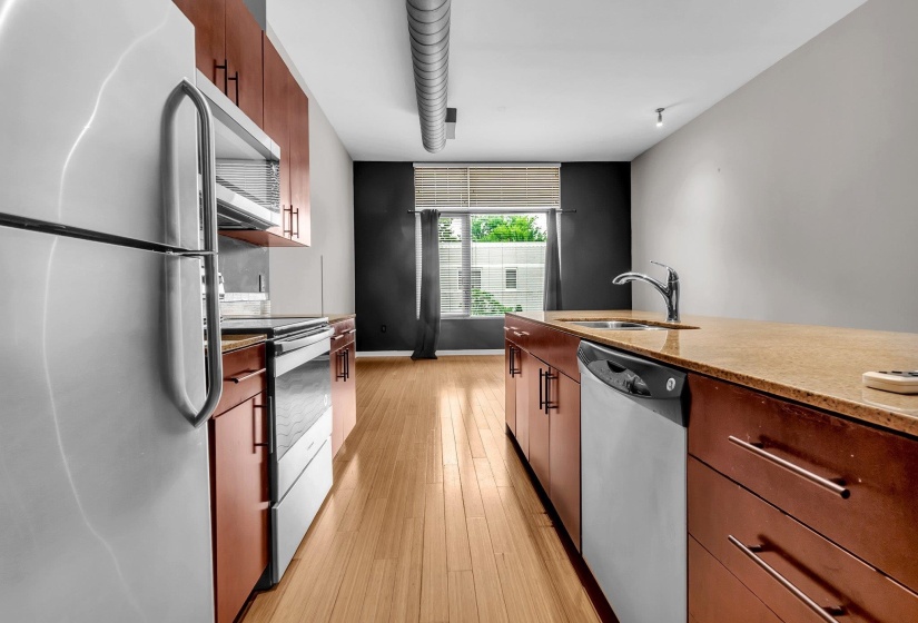 Kitchen featuring wood-finish flooring, stainless steel appliances, wood cabinetry with modern hardware, stone countertops, and a recessed sink with a chrome faucet