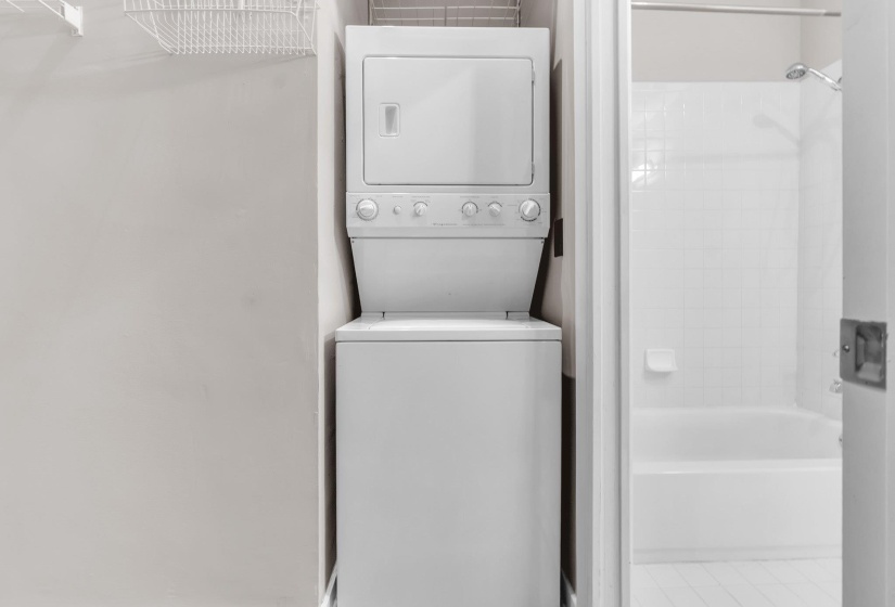 Compact laundry area featuring a white stackable washer and dryer unit, wire shelving, and a nearby bathroom with a white bathtub and tiled shower surround
