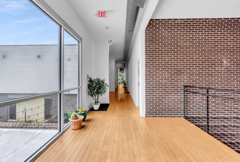 Hallway featuring wood-finish flooring, a full-height window, exposed brick accent wall, and contemporary black metal railings