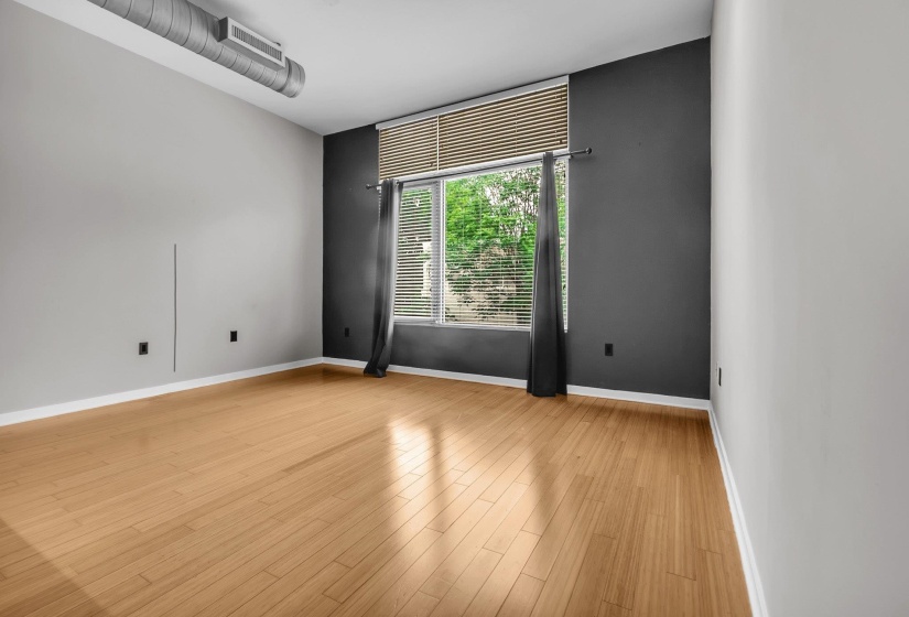 Spacious room featuring wood-finish flooring, a large window with blinds, and an exposed industrial-style duct