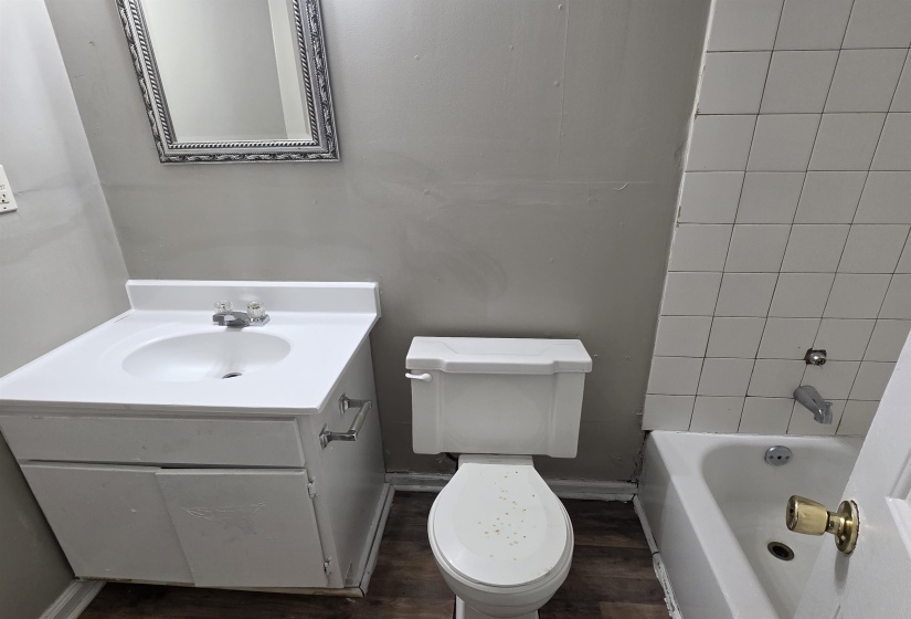Bathroom featuring a white vanity with integrated sink, chrome faucet, white toilet, white bathtub with white tiled surround, and wood-finish flooring