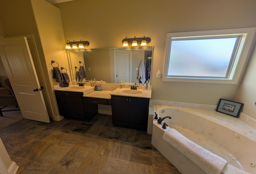 Bathroom featuring a corner jetted soaking tub, dual sink vanity with stone-finish countertops, tiled flooring, and a frosted window