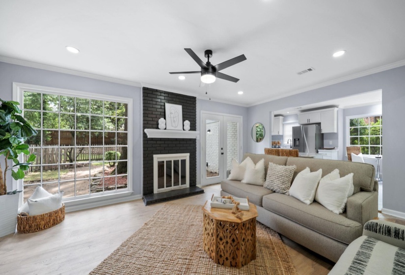 Spacious living area featuring a large grid-pattern window, a dark brick fireplace with white mantel, a ceiling fan with integrated lighting, recessed lighting, and light wood-finish flooring