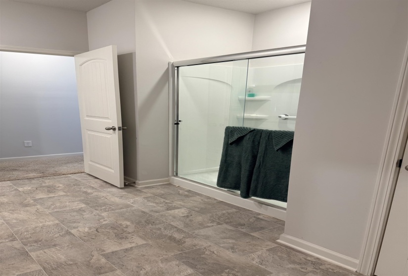 Spacious bathroom featuring a glass-enclosed shower with integrated shelving, a white arched panel door, light gray wall paint, white baseboards, and stone-look tile flooring