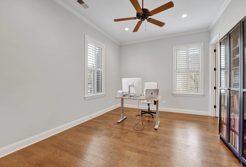 Room featuring wood-finish flooring, two windows with plantation shutters, crown molding, and a ceiling fan with integrated lighting