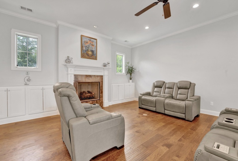 Living room featuring wood-finish flooring, a fireplace with a stone surround and white mantel, built-in lower cabinetry with white paneled doors, white crown molding, and recessed lighting