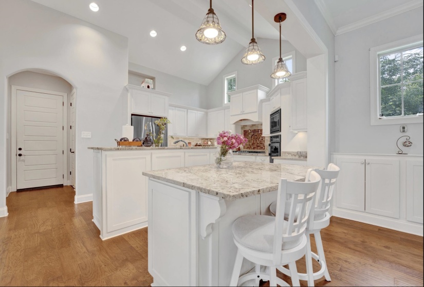 Spacious kitchen featuring an expansive island with a light-toned countertop, white cabinetry, integrated stainless-steel appliances, recessed lighting, and a vaulted ceiling