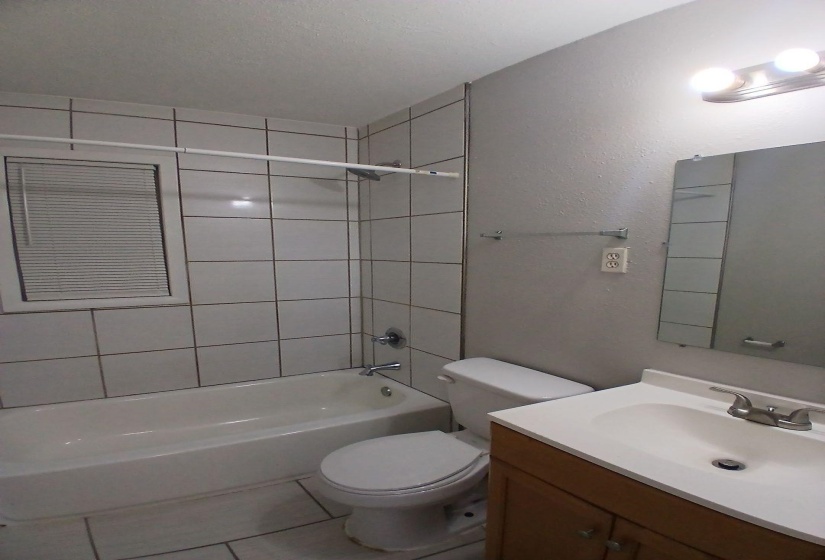 Bathroom featuring a white subway-tiled tub/shower combination, window with blinds, wood-finish vanity with an integrated sink, and wall-mounted mirror