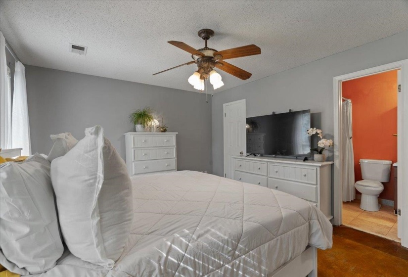 Bedroom featuring a ceiling fan with light fixture, light gray walls, and an open doorway revealing a bathroom with a toilet and shower curtain